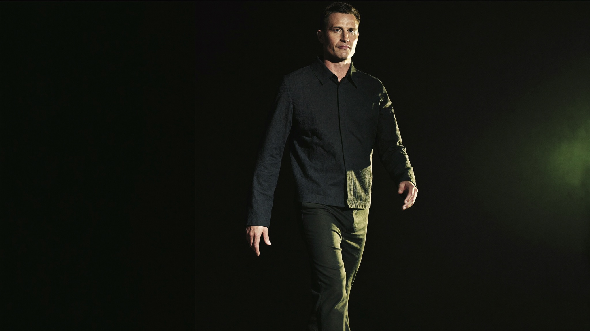 A self-possessed adult walking forward with quiet strength years after a major weight-loss journey, fully clothed, dark editorial setting